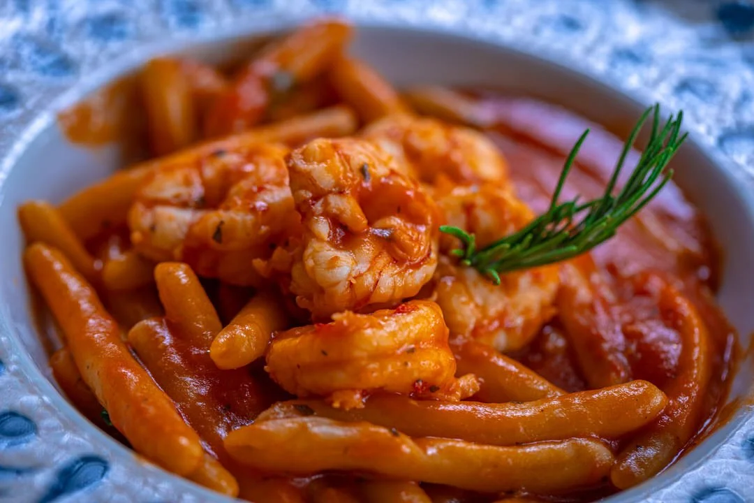Close-up of a bowl of shrimp and pasta with tomato sauce garnished with a sprig of rosemary.