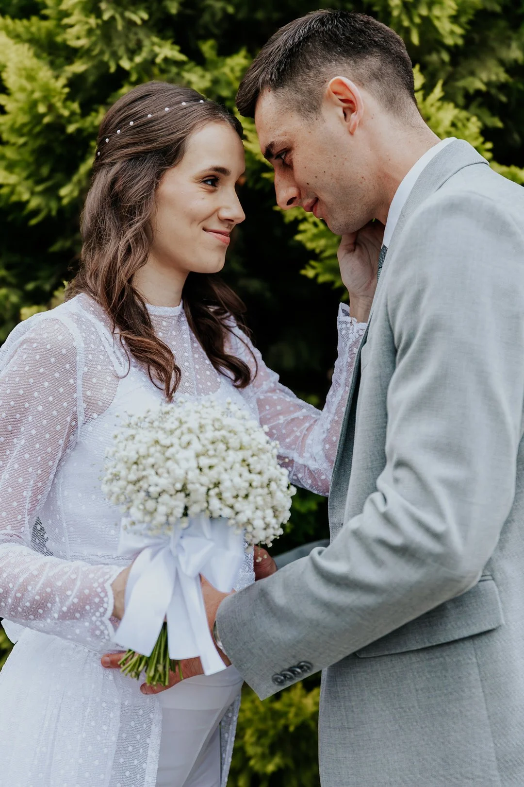 A bride and groom face each other outdoors, the bride holding a white flower bouquet, both smiling softly, surrounded by greenery.