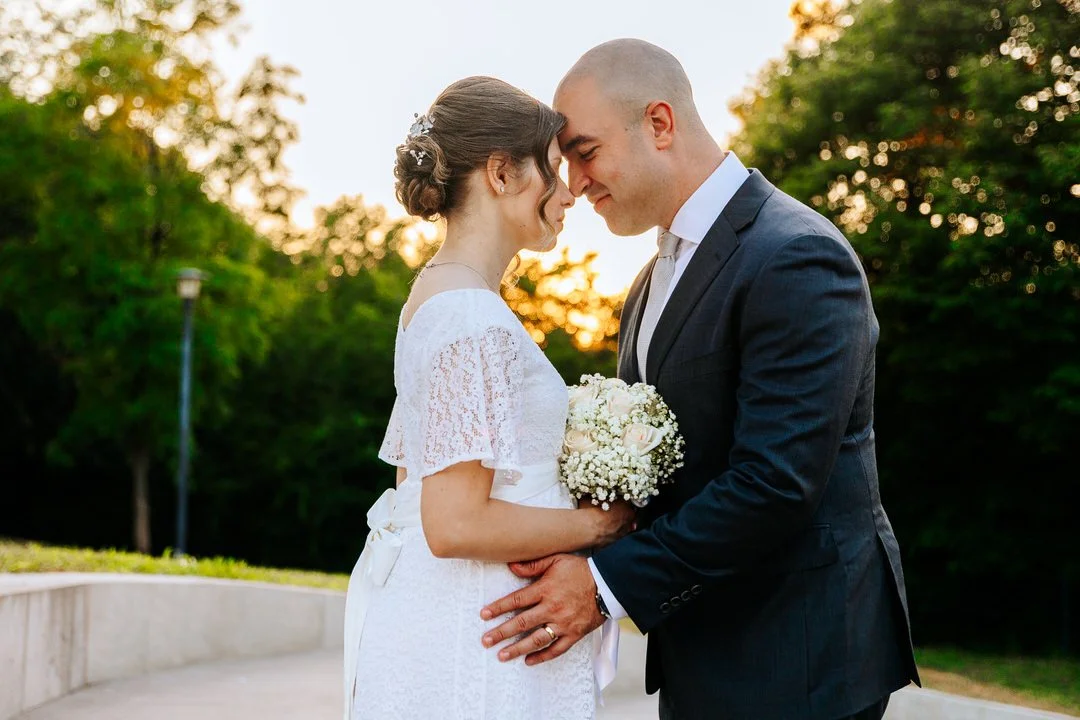 A bride and groom stand close together with foreheads touching outdoors during sunset, with green trees in the background. The bride holds a bouquet of white flowers, and both are smiling softly.