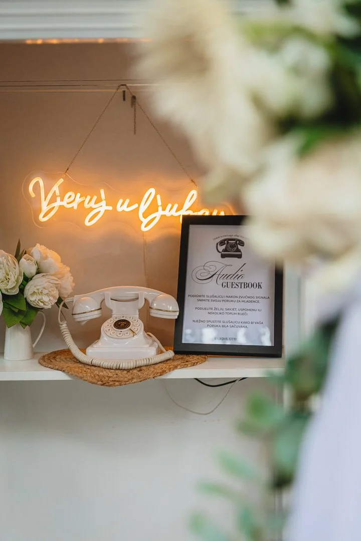 Decorative reception area with a vintage white rotary telephone on a small woven mat, framed guestbook sign, and a neon sign in a script font that reads 'Vjeruj u ljubav.'