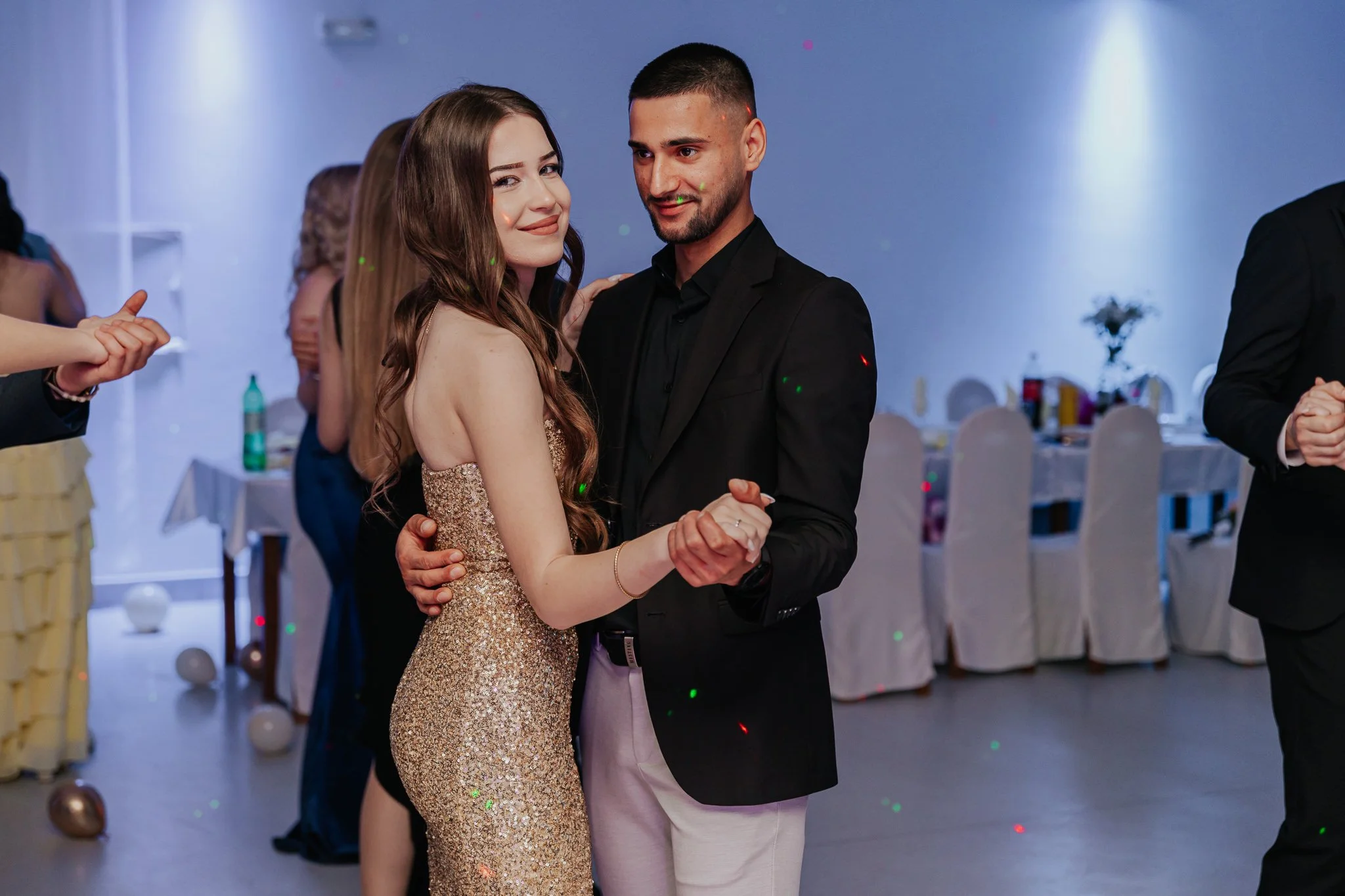 A couple dancing together at a formal event, with the woman wearing a gold dress and the man in a black suit.
