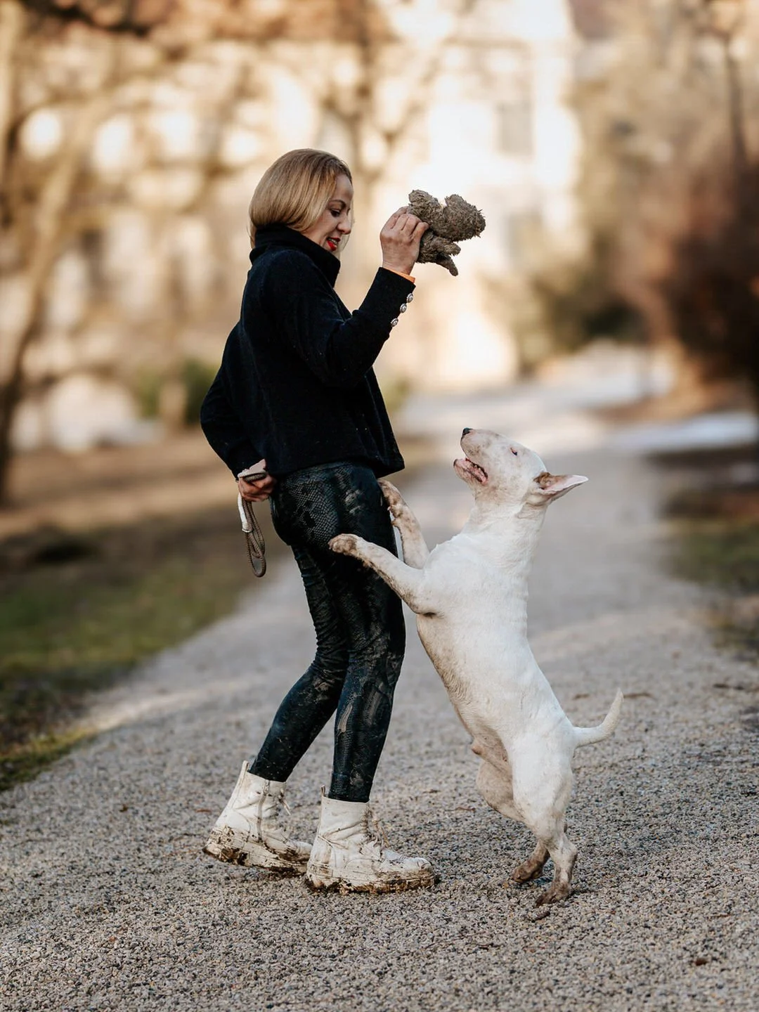 A woman holding a toy in the air while a dog jumps up to her, standing on its hind legs with front paws on her. They are outdoors on a gravel path with trees and buildings in the background.