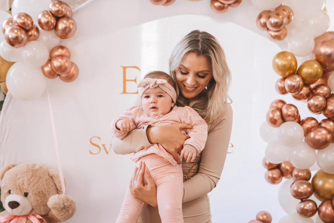 Woman smiling and holding a baby girl surrounded by balloon decorations at a celebration following a Croatia baptism in Ložiona Orahovica, including white, rose gold, and gold balloons.