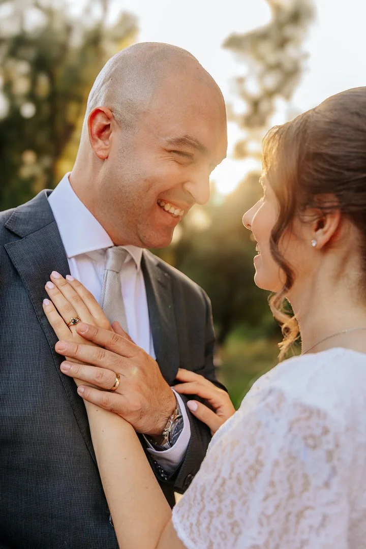 A newlywed couple smiling and looking at each other outdoors during sunset, holding hands and embracing.
