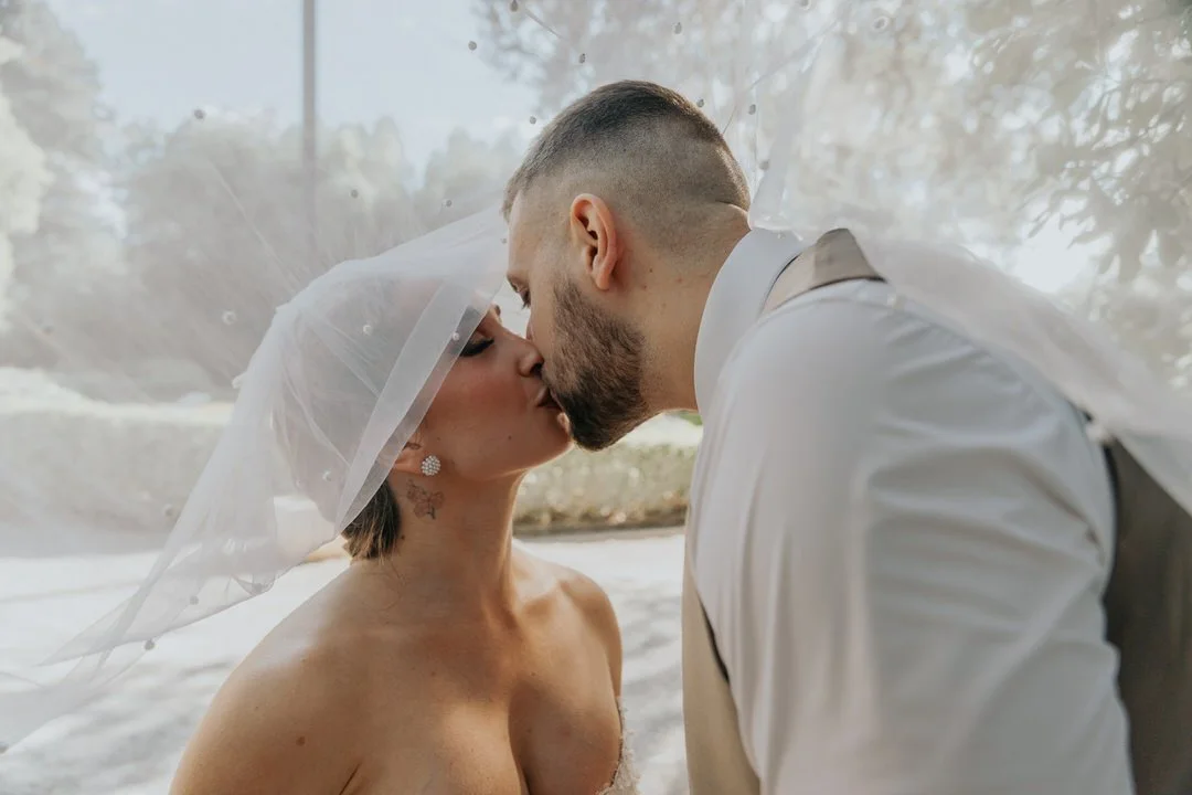 A couple dressed in wedding attire sharing a kiss outside, with both wearing white. The bride has tattoos and a veil, and the groom has a beard and short hair.