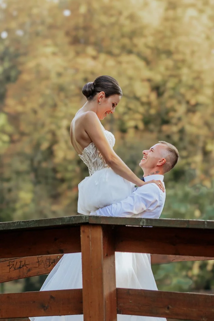 A bride and groom in wedding attire on a wooden platform outdoors with autumn trees in the background. The groom lifts the bride who is smiling and looking down at him.
