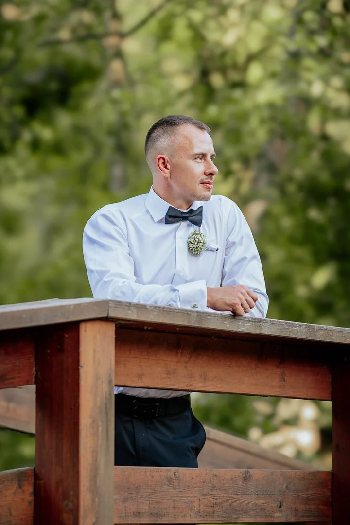 A groom dressed in a white shirt with a black bow tie and boutonniere, standing on a wooden balcony outdoors with green trees in the background.