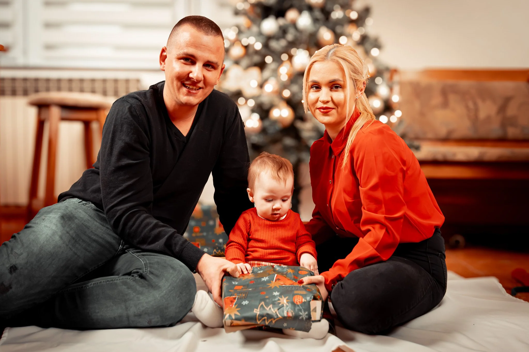 A family of three, including a man, a woman, and a young child, sitting on the floor in front of a decorated Christmas tree, opening a wrapped gift together, with a festive background.