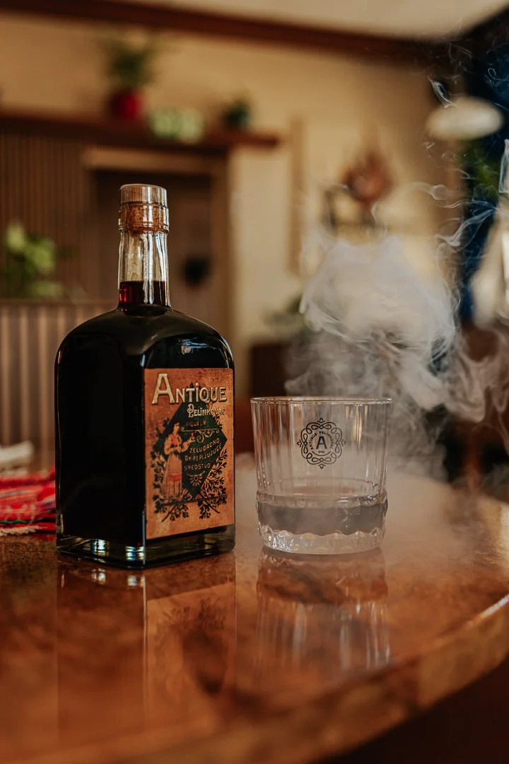 A bottle of Antique Pelinkovac with a dark liquid inside, next to a glass with some smoke rising from it, on a wooden surface in a cozy Roots & Flames restaurant Bobingen