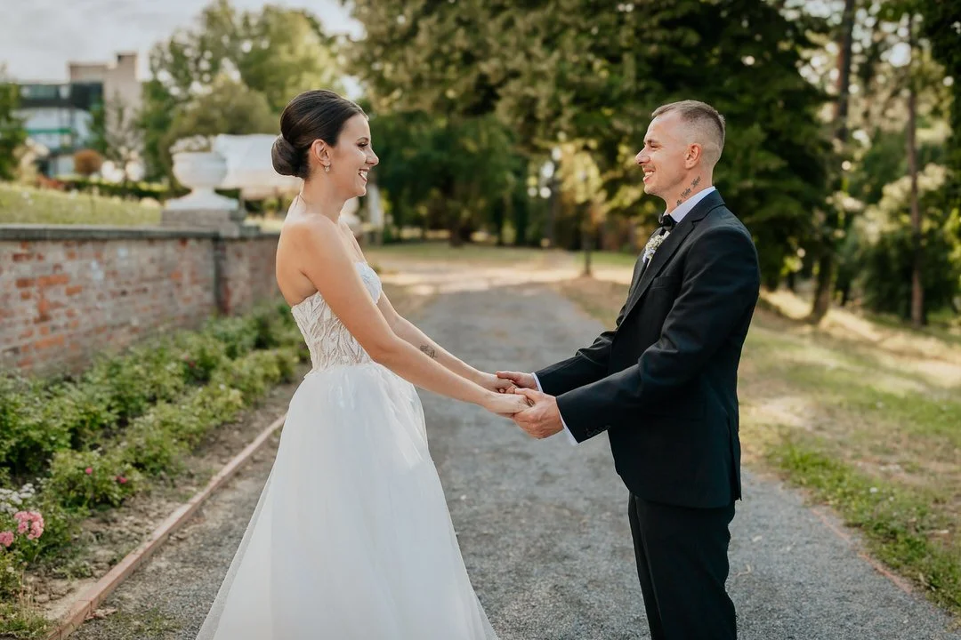 A bride and groom holding hands and smiling at each other outdoors with trees and a brick wall in the background.
