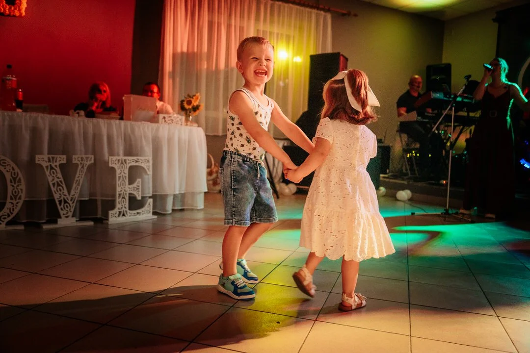 Two children, a boy and a girl, are dancing together at a celebration, with the boy smiling and the girl wearing a white dress with a large bow in her hair.