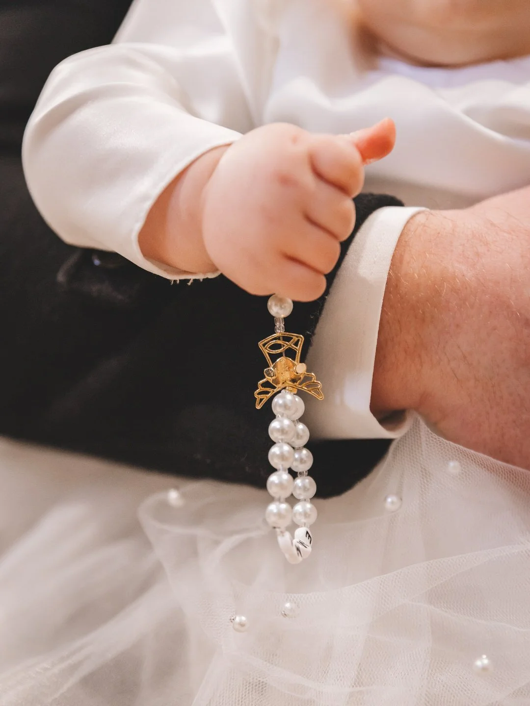 Close-up of a child's hand holding a pearl beaded rosary with a gold angel charm during a Croatia baptism in Slavonija, against a background of a white satin garment and tulle fabric.