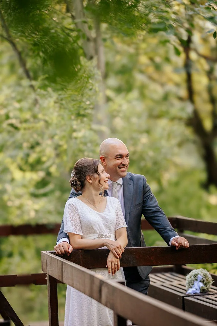 A bride and groom standing outdoors on a wooden bridge in park forest Adica in Vukovar, smiling and looking at each other with trees in the background.