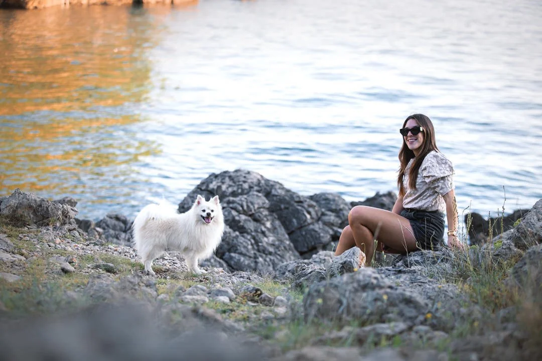 A woman sitting on rocks by a body of water, wearing sunglasses and smiling, with a fluffy white dog beside her in Prniba, Krk Island, Croatia.