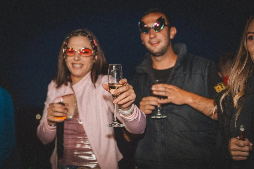 Three young adults at a 1990's birthday party holding glasses of champagne, wearing sunglasses, smiling, at night
