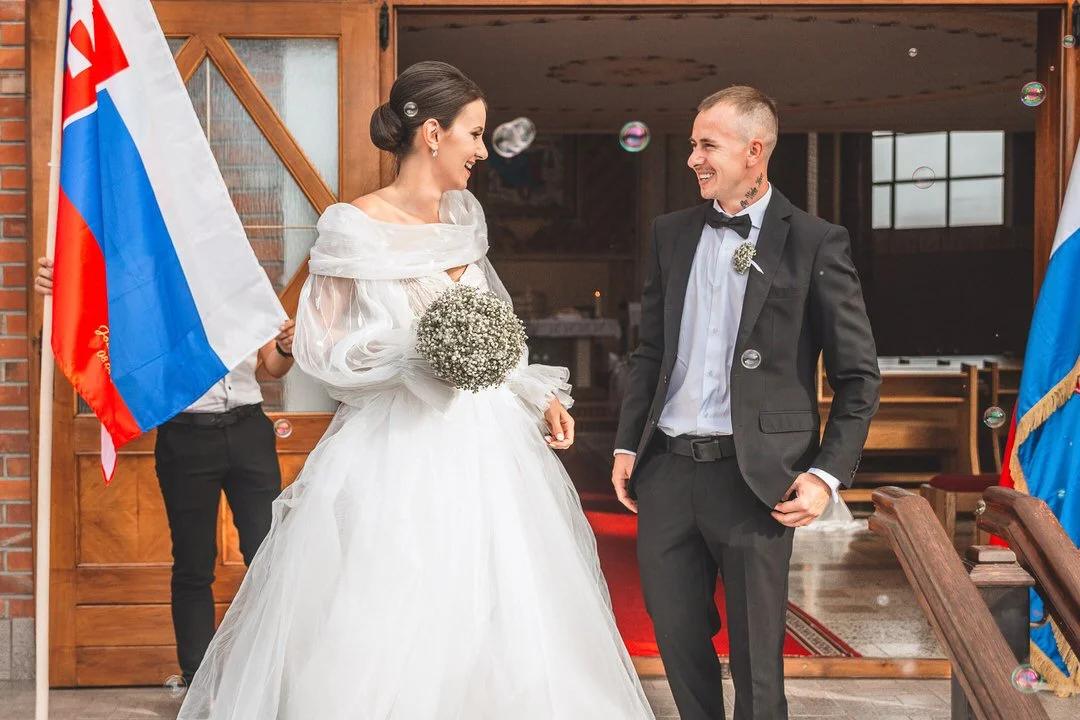 A bride and groom smiling at each other during their wedding ceremony, with a person holding a Russian flag on either side of them.