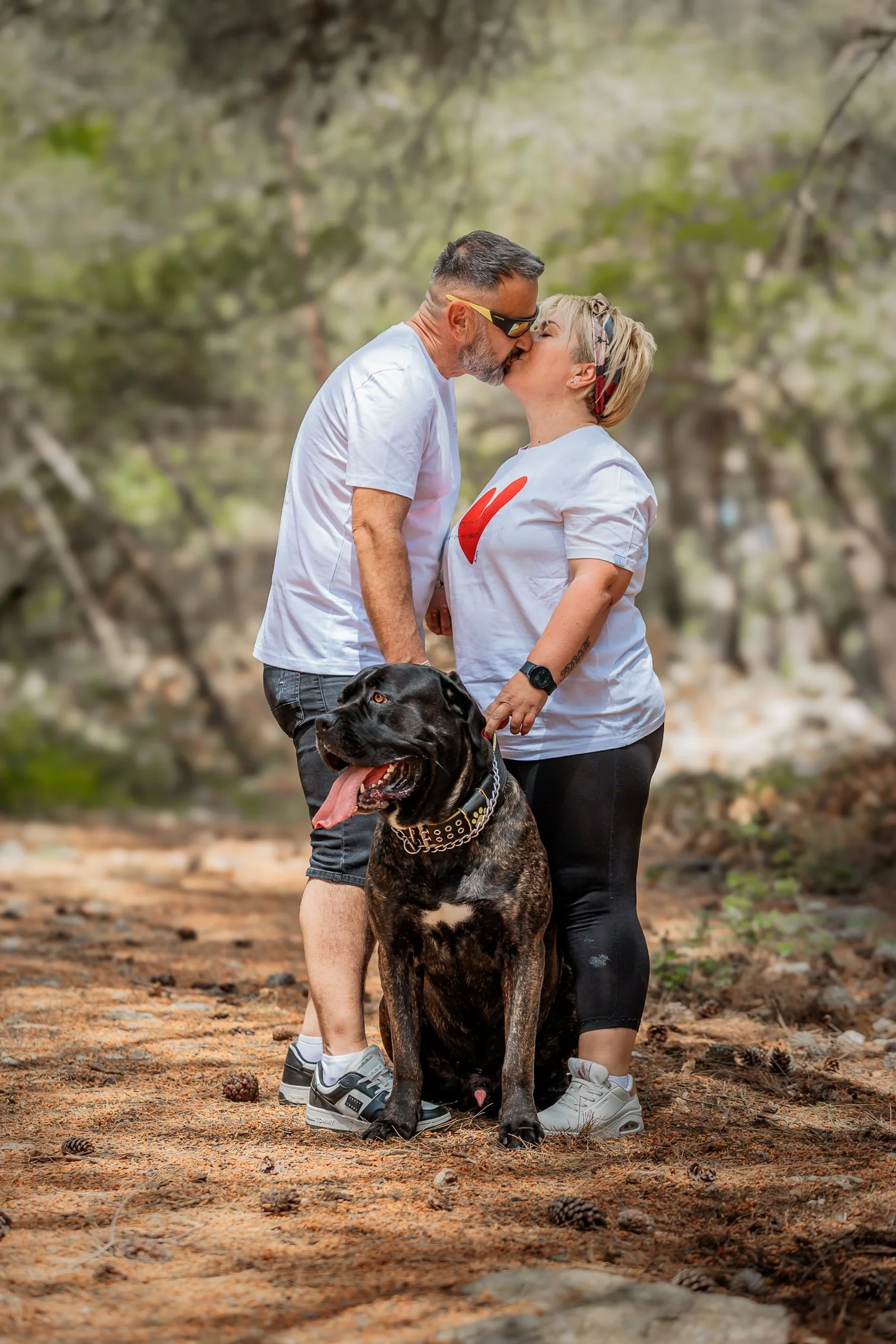 A couple kissing outdoors on a nature trail with a cane corso dog between them, surrounded by trees and fallen leaves on Krk Island Croatia