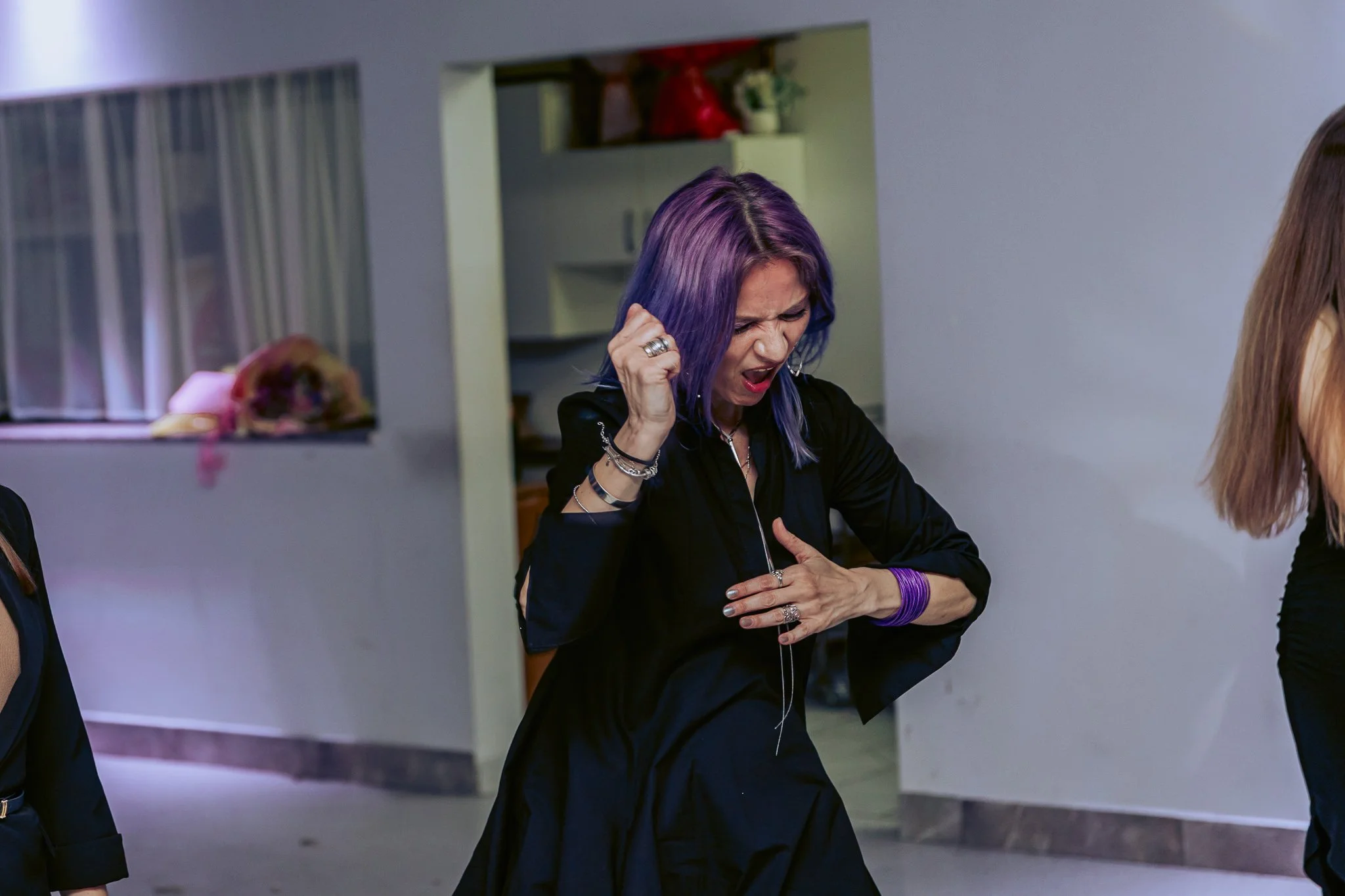 A woman with vibrant purple and blue hair, wearing jewelry, dancing at the party . She is indoors, and part of another person is visible on the right side of the image.