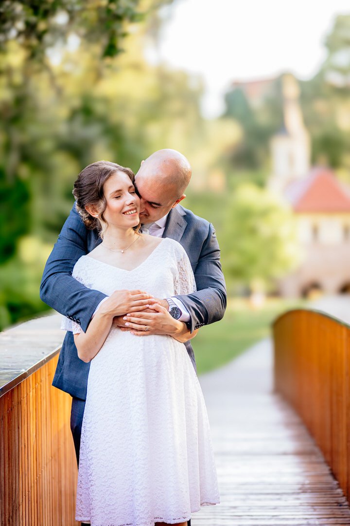A couple in wedding attire embracing on a wooden bridge by a river with a blurred scenic background of trees and buildings.