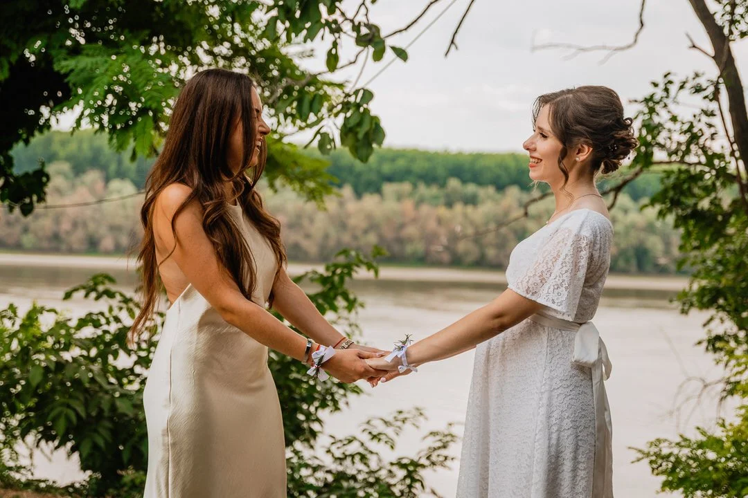 Two women stand by a river, holding hands and smiling at each other during a wedding or commitment ceremony, surrounded by greenery.