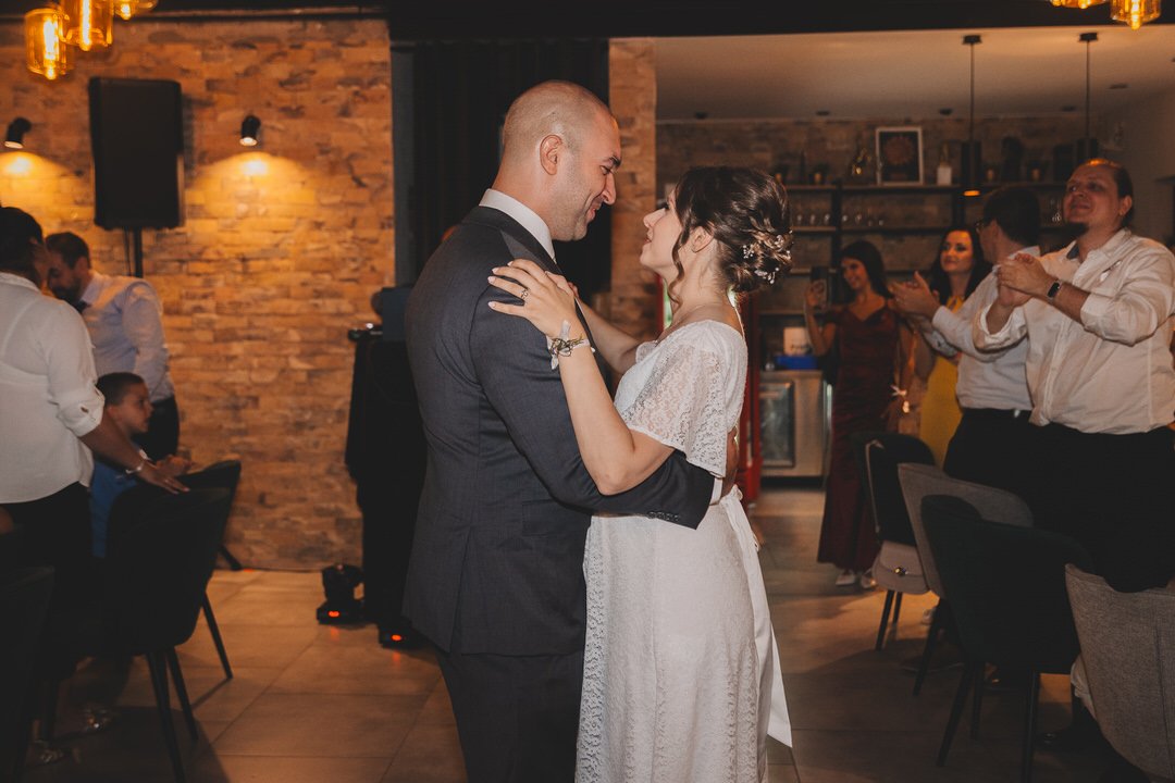 Bride and groom dancing at a wedding reception at Dunavski Raj wedding venue, surrounded by seated and standing guests in a warmly lit indoor space with brick walls.