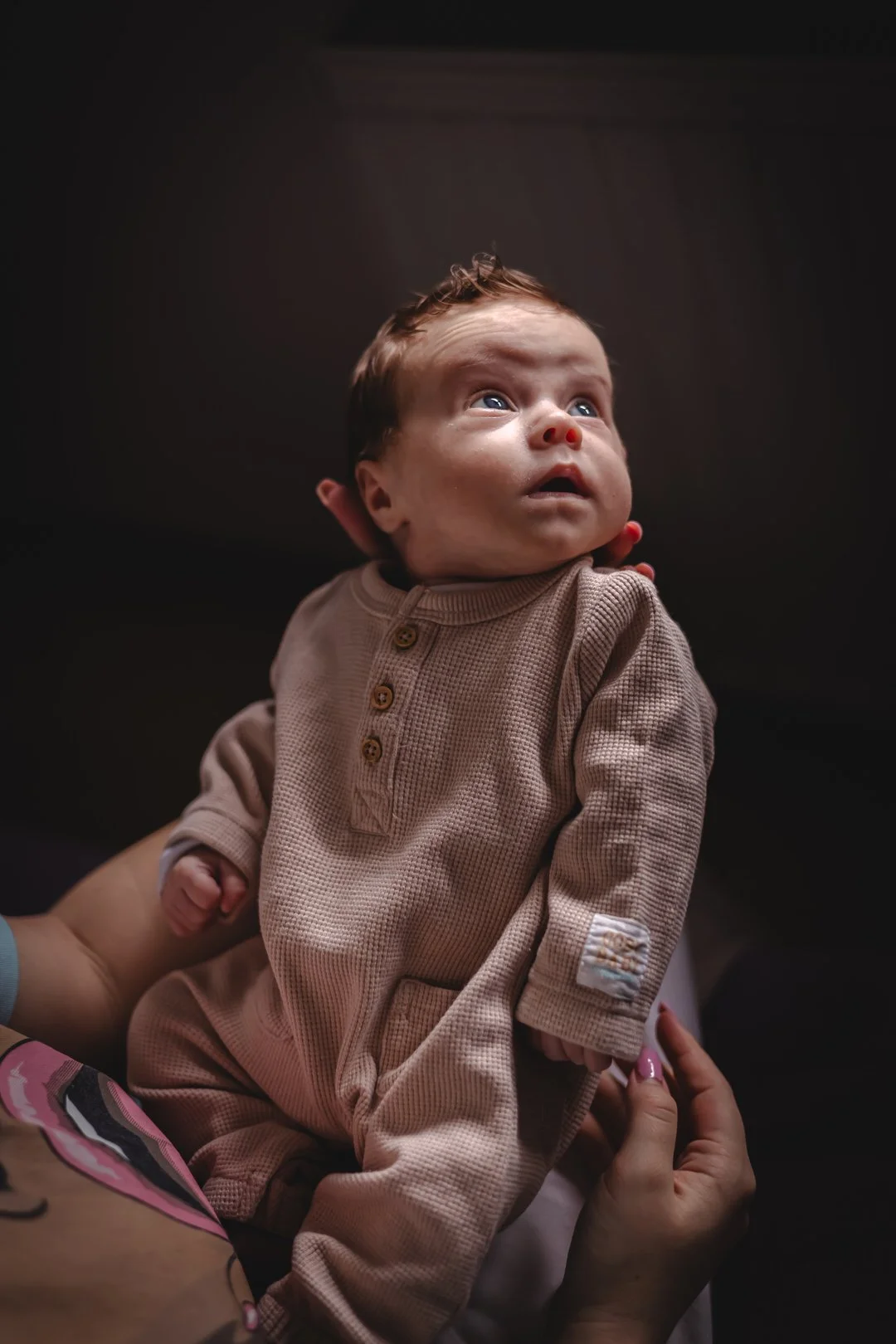 A baby with a reddish nose and short brown hair looking upwards, wearing a beige textured outfit with buttons, being held by someone with pink nail polish.