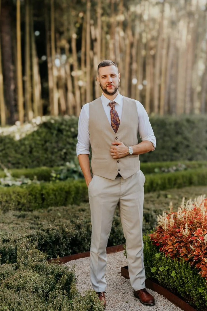 Groom wearing a beige vest, white shirt and paisley tie standing in Park Angiolina, Opatija, Kvarner, surrounded by lush garden greenery.