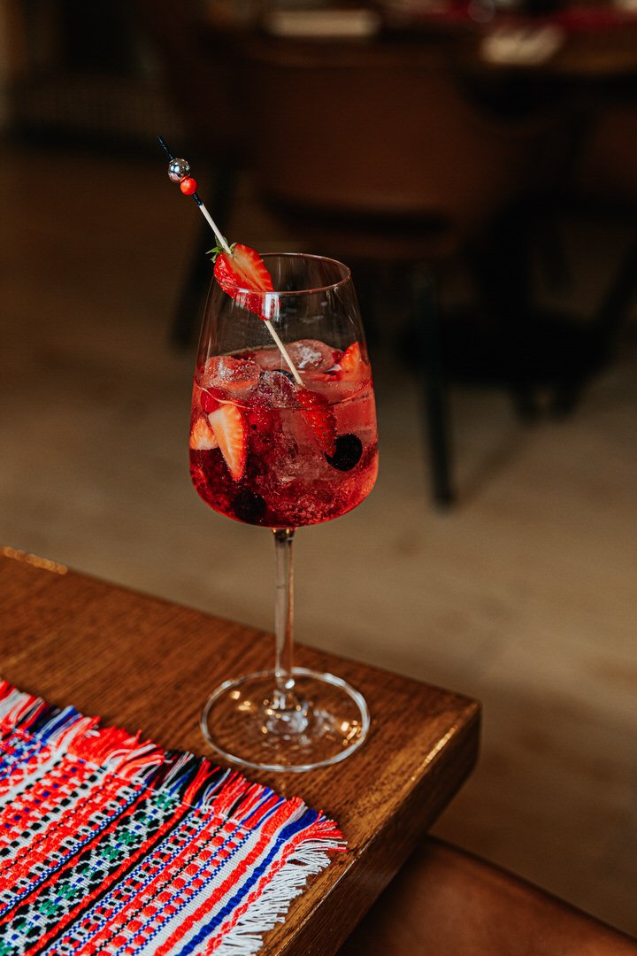 A glass of red mixed drink with strawberries and blueberries, garnished with a strawberry slice and a cocktail pick, on a wooden table with a colorful woven cloth.