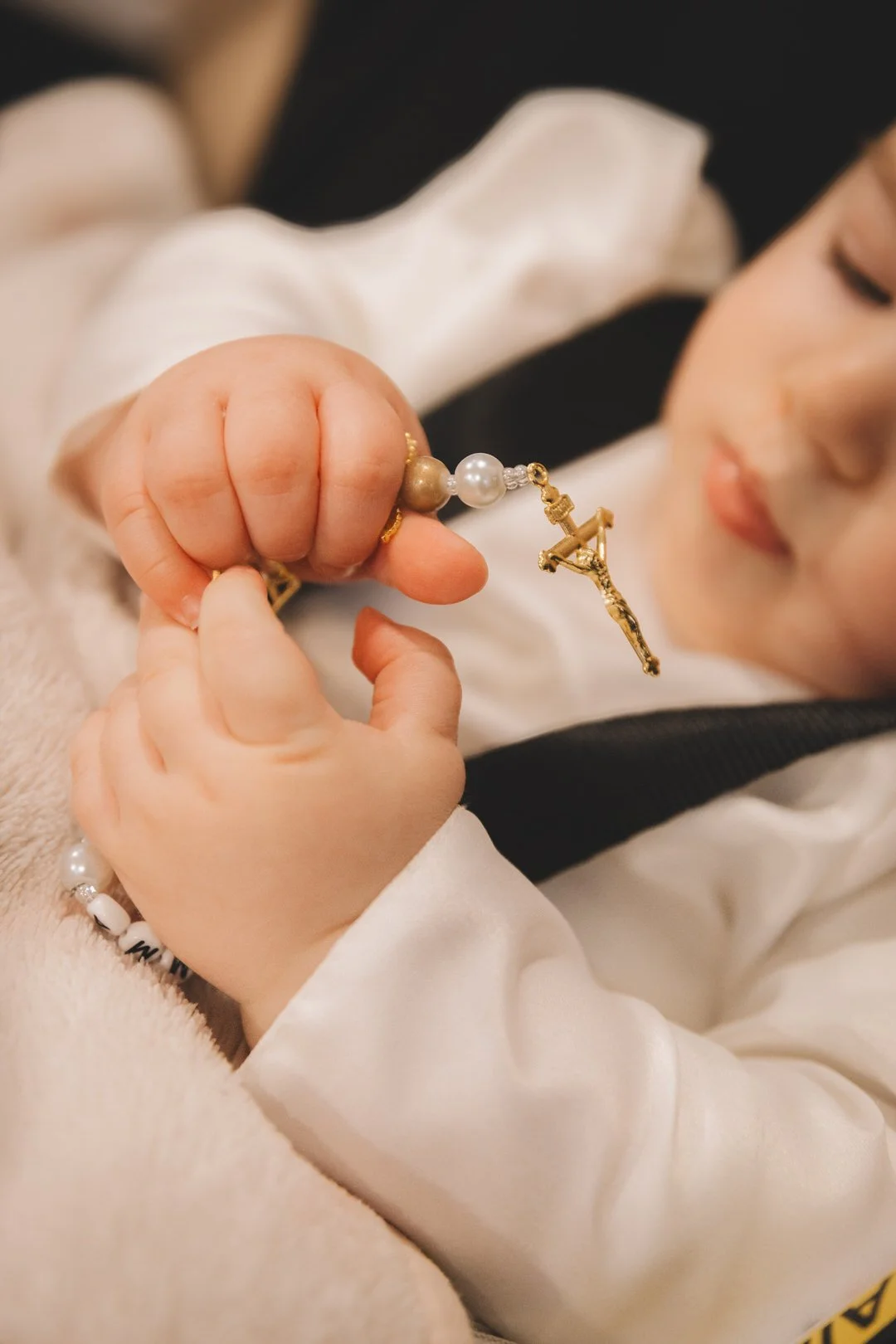 A young child holding a gold rosary with a crucifix , the child's hand and face are visible, and they are wearing a white garment.
