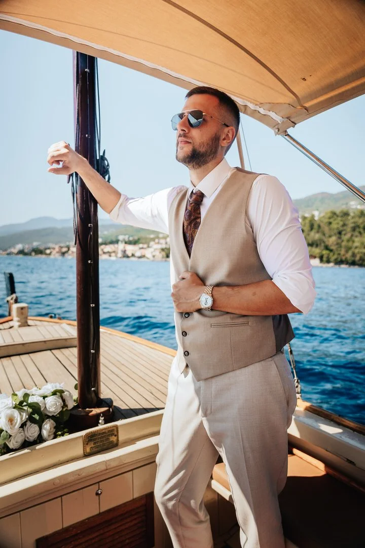 A man wearing sunglasses, a beige vest, white shirt, and beige pants standing on a boat with water and a distant shoreline in the background.