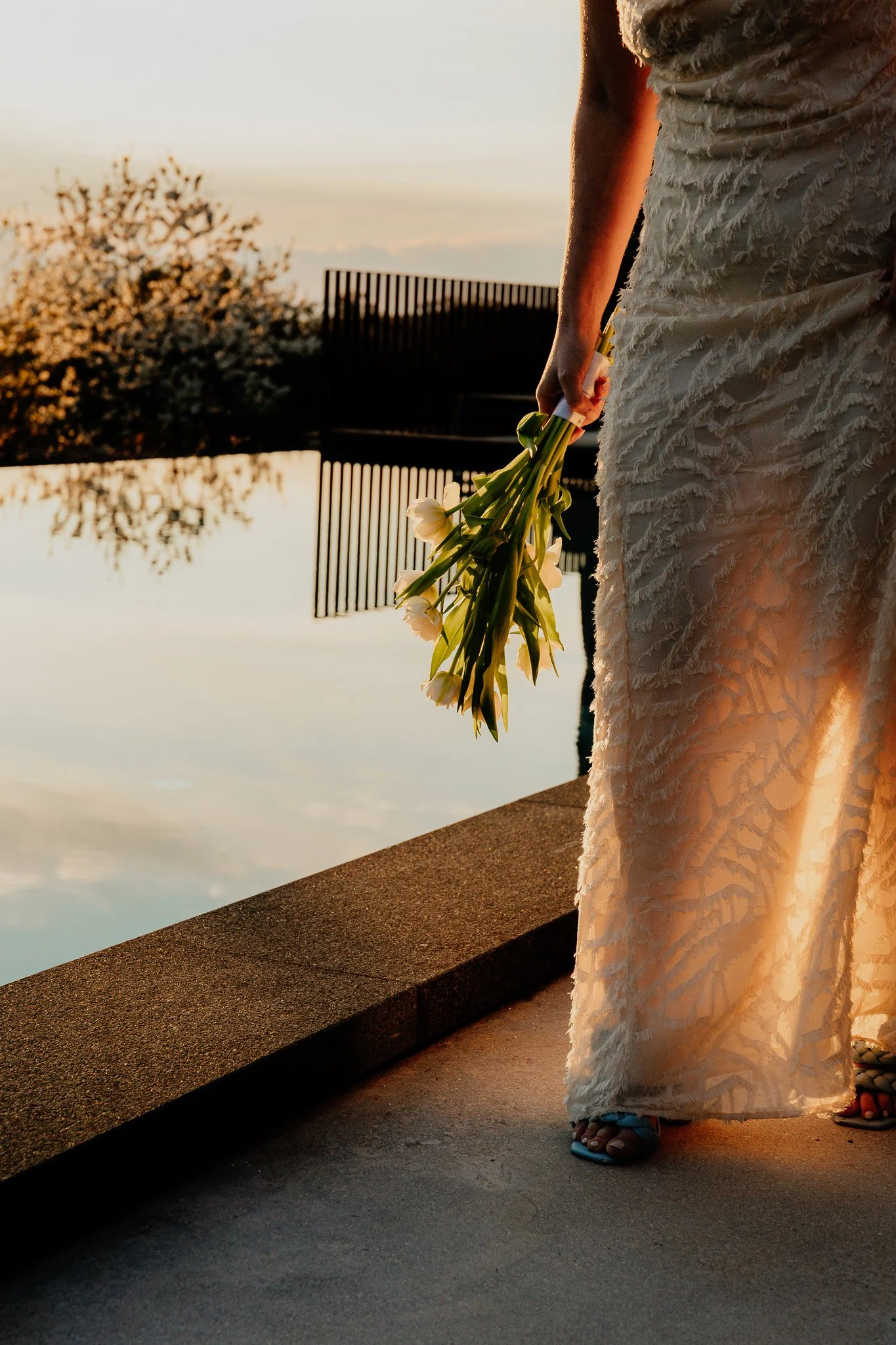 a bride wearing a textured beige dress holding white flowers and standing near water at sunset.