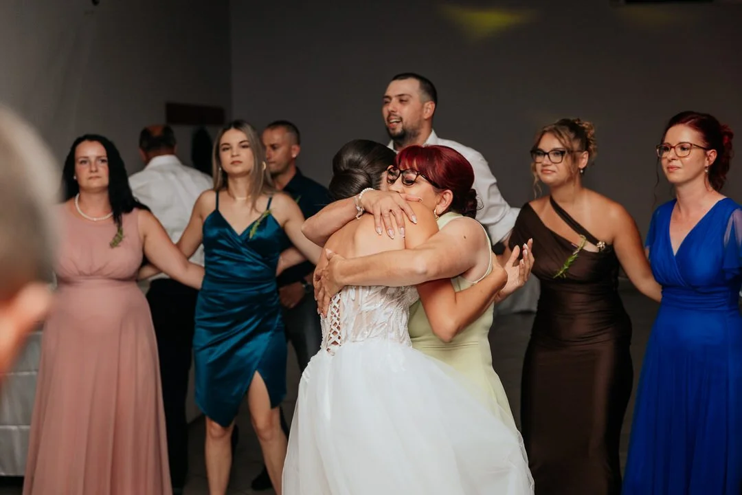 People dancing and hugging at a wedding reception, with women in colorful dresses and a man in a white shirt in the background.