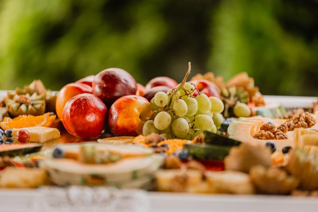Close-up of a fruit and cheese platter with grapes, apples, blueberries, and various cheeses and nuts, outdoors with a blurred green background.