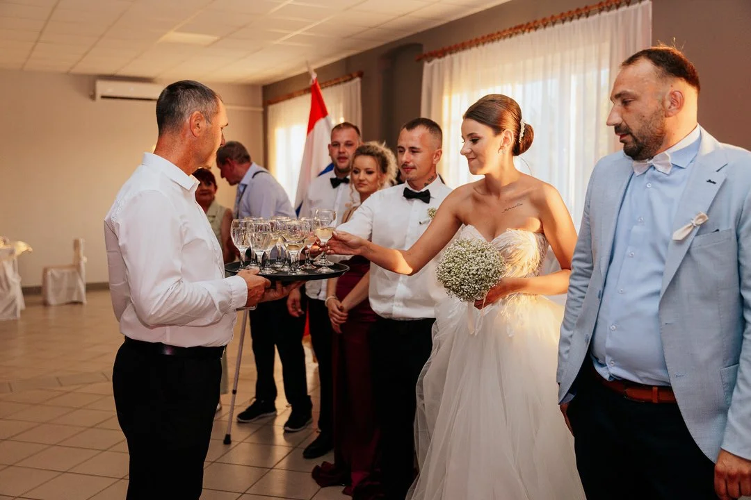 Bride in a wedding dress holding a bouquet and receiving drinks from a server, with friends and family in formal attire standing nearby during a wedding celebration.