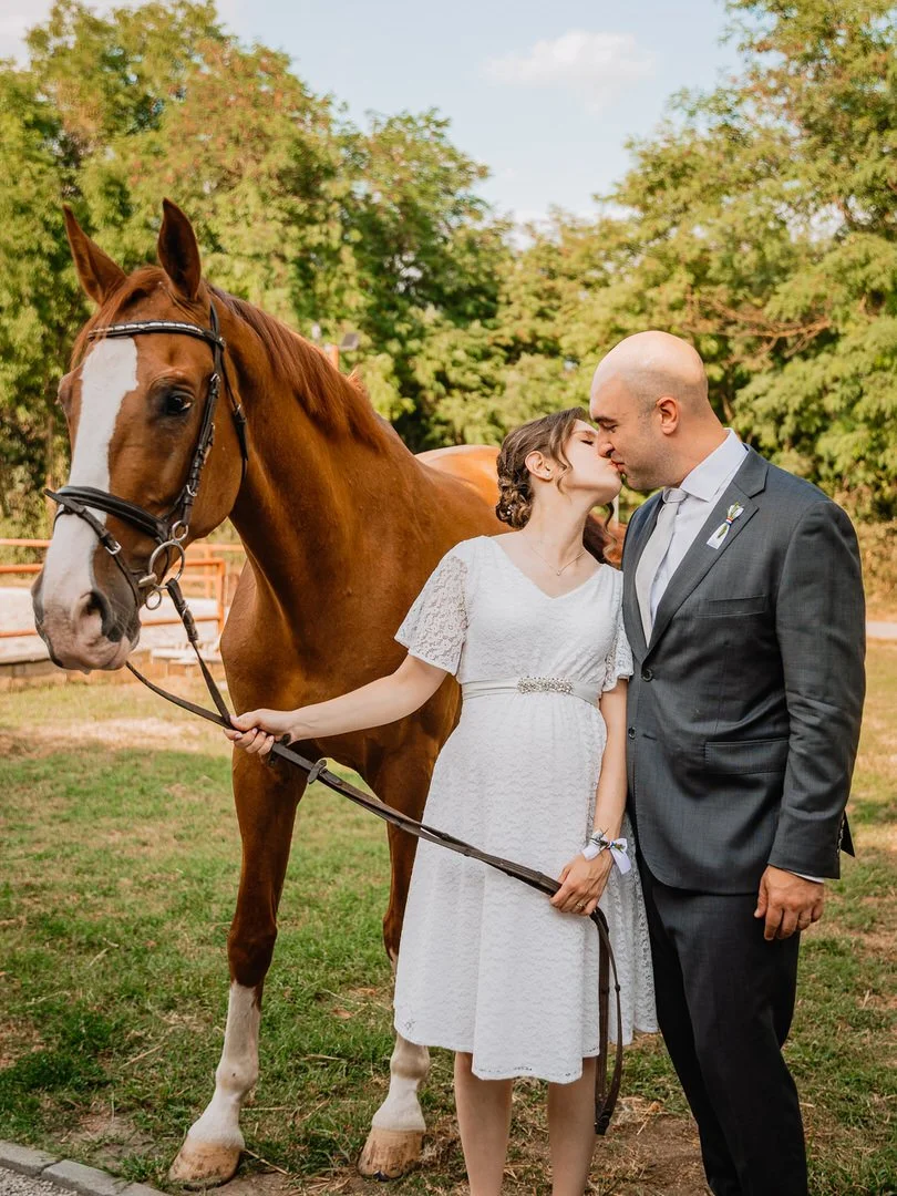 A bride and groom standing in a park, about to kiss, with a brown horse nearby. The bride is in a white dress and the groom in a gray suit. The horse is on a leash held by the bride, and there are trees in the background.