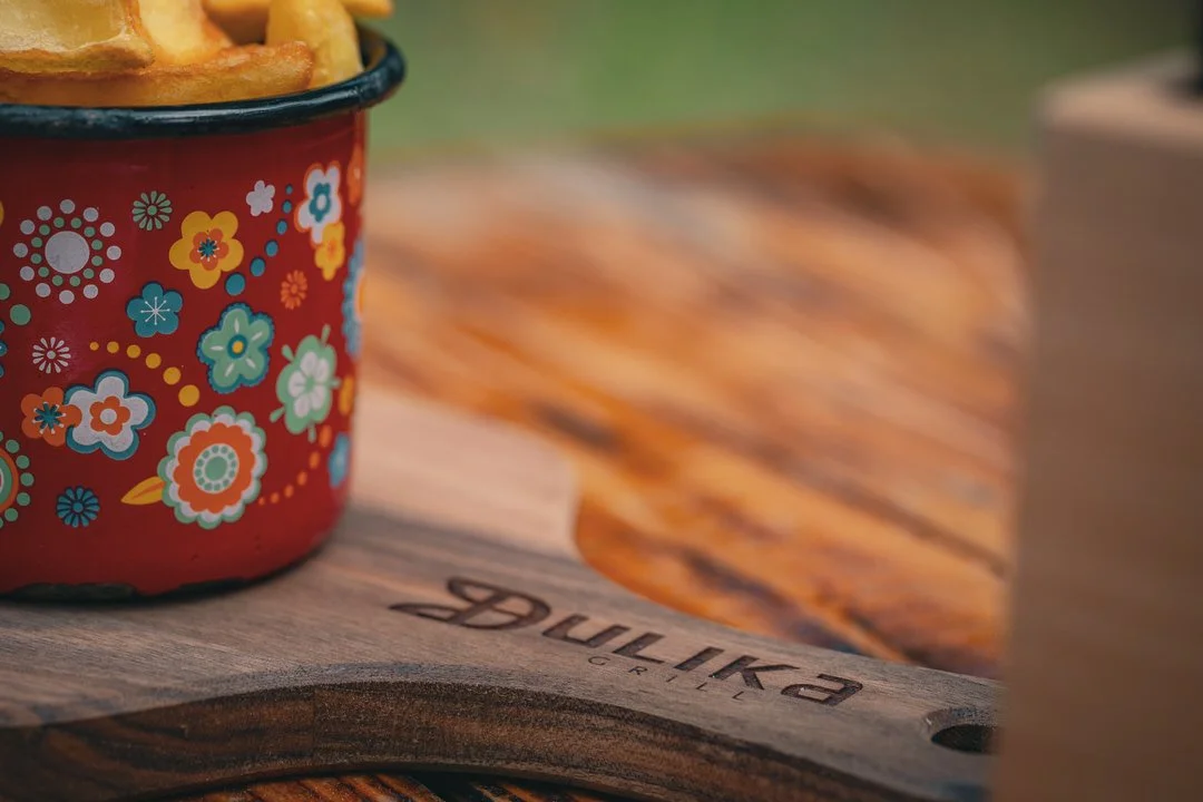 Close-up of a red enamel mug with colorful floral patterns, filled with French fries, placed on a wooden cutting board outdoors in Ulika grill restaurant in Vrbnik Croatia