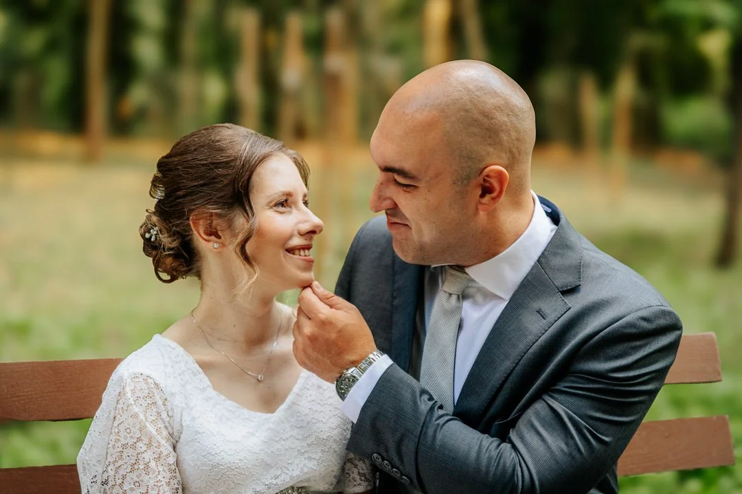 A man in a gray suit and a woman in a white dress sitting together on a wooden bench outdoors, gazing at each other lovingly with blurred trees in the background.