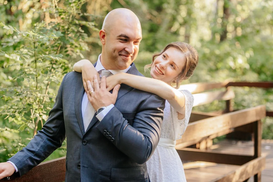 A man and woman, smiling and enjoying each other's company outdoors on a wooden bridge surrounded by green trees.