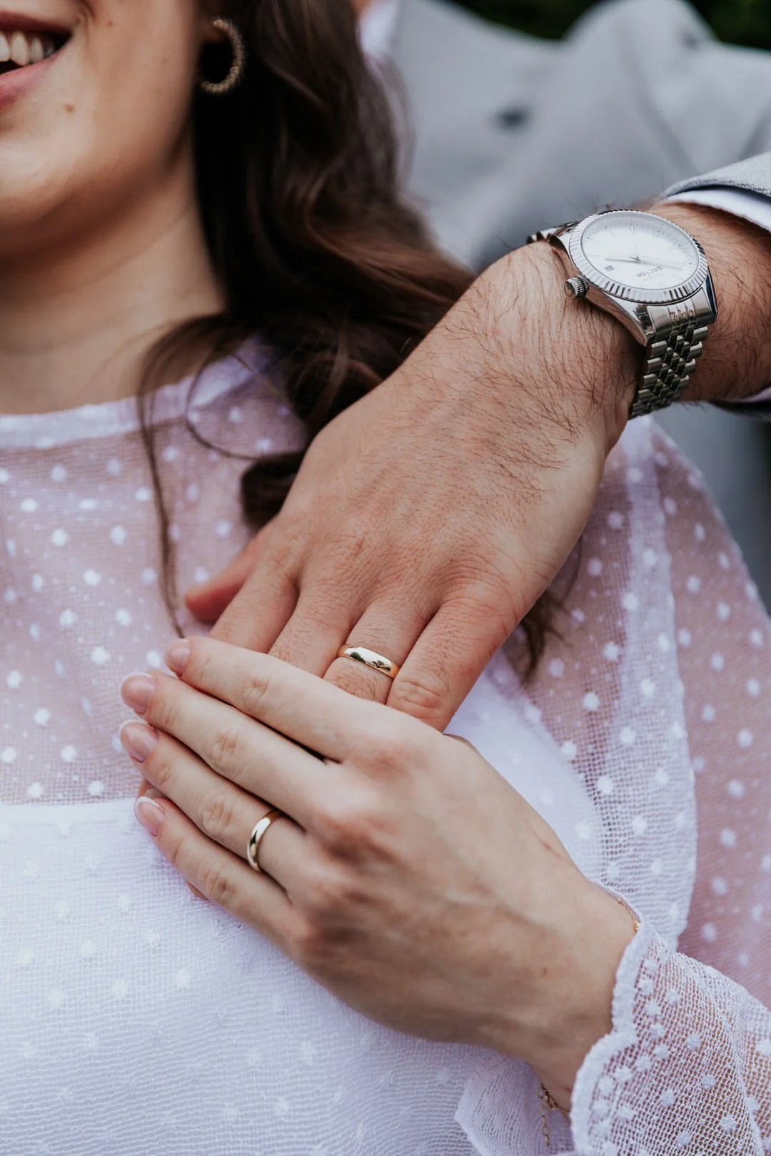 Close-up of a couple's hands, one with a wedding band and the other with a watch, resting on a woman's shoulder, with her wearing a white and pink polka dot top.