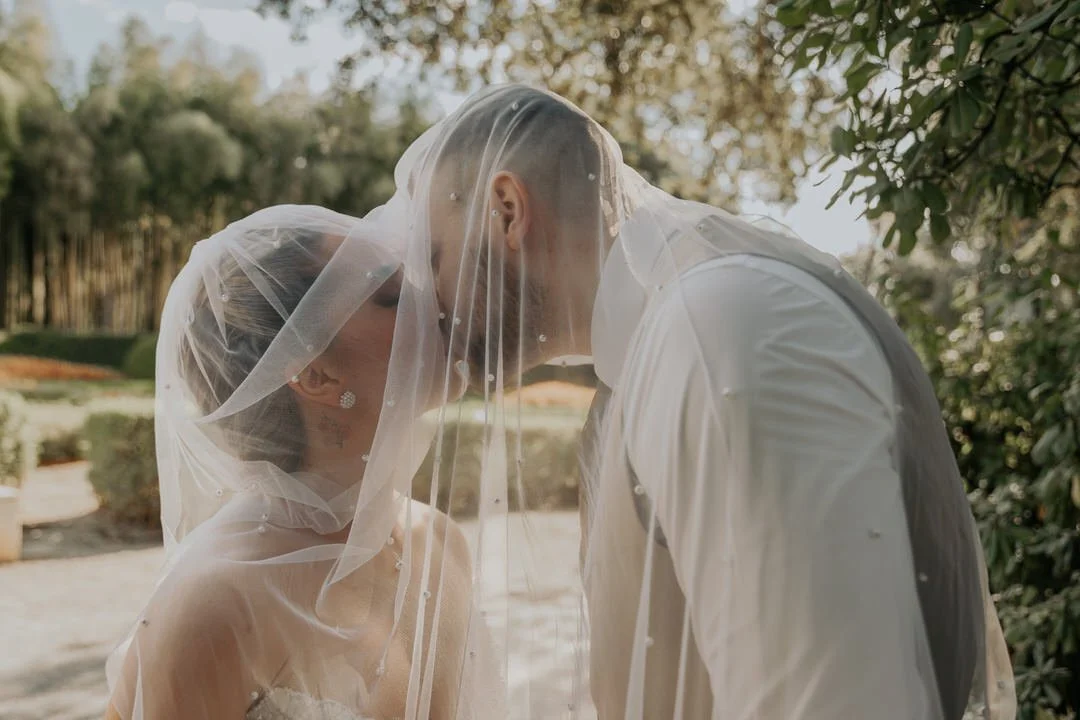 Bride and groom standing closely together under a veil outdoors.