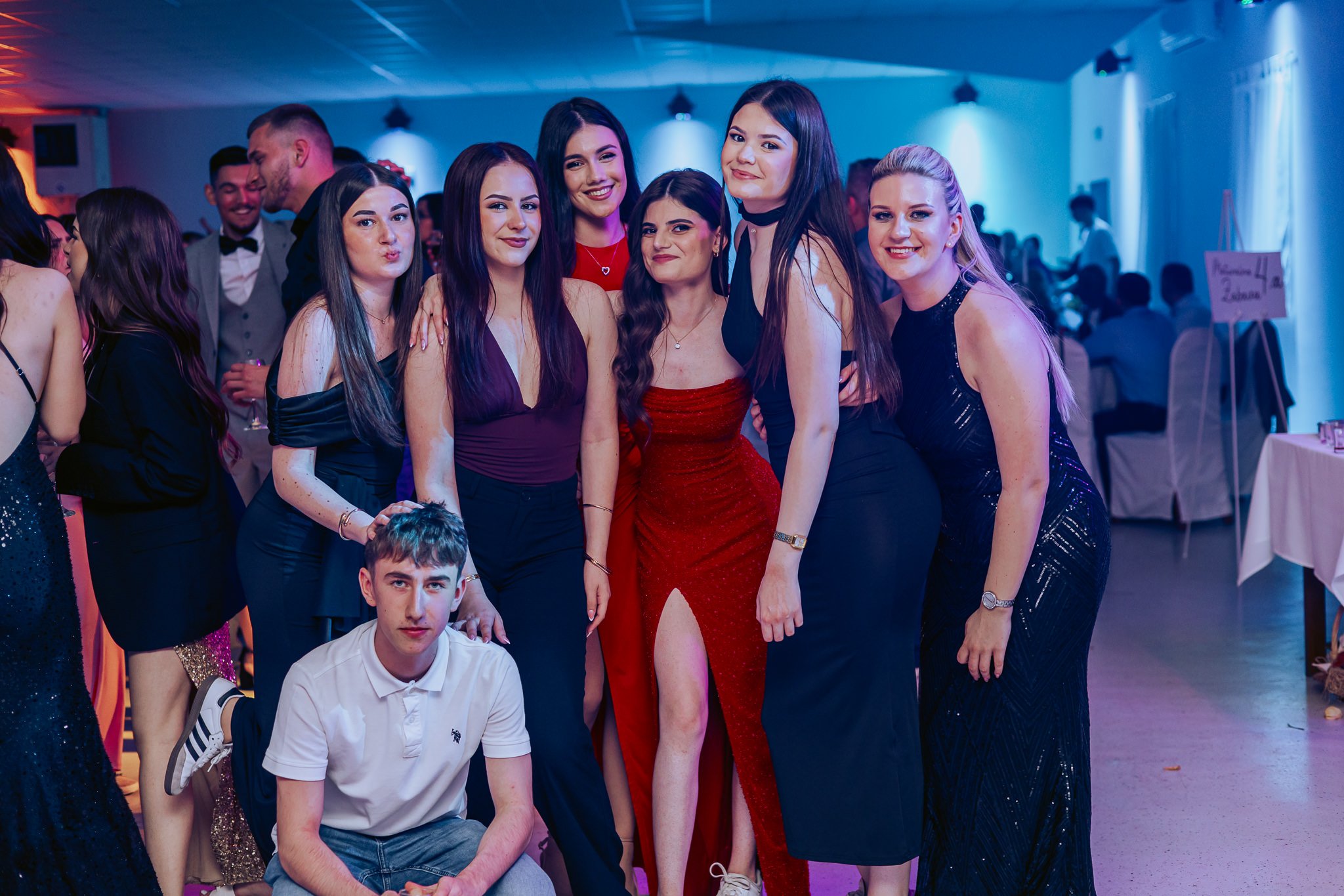 A group of young people, mostly women in formal dresses, posing and smiling at a prom party or celebration in an indoor venue with colorful lighting.
