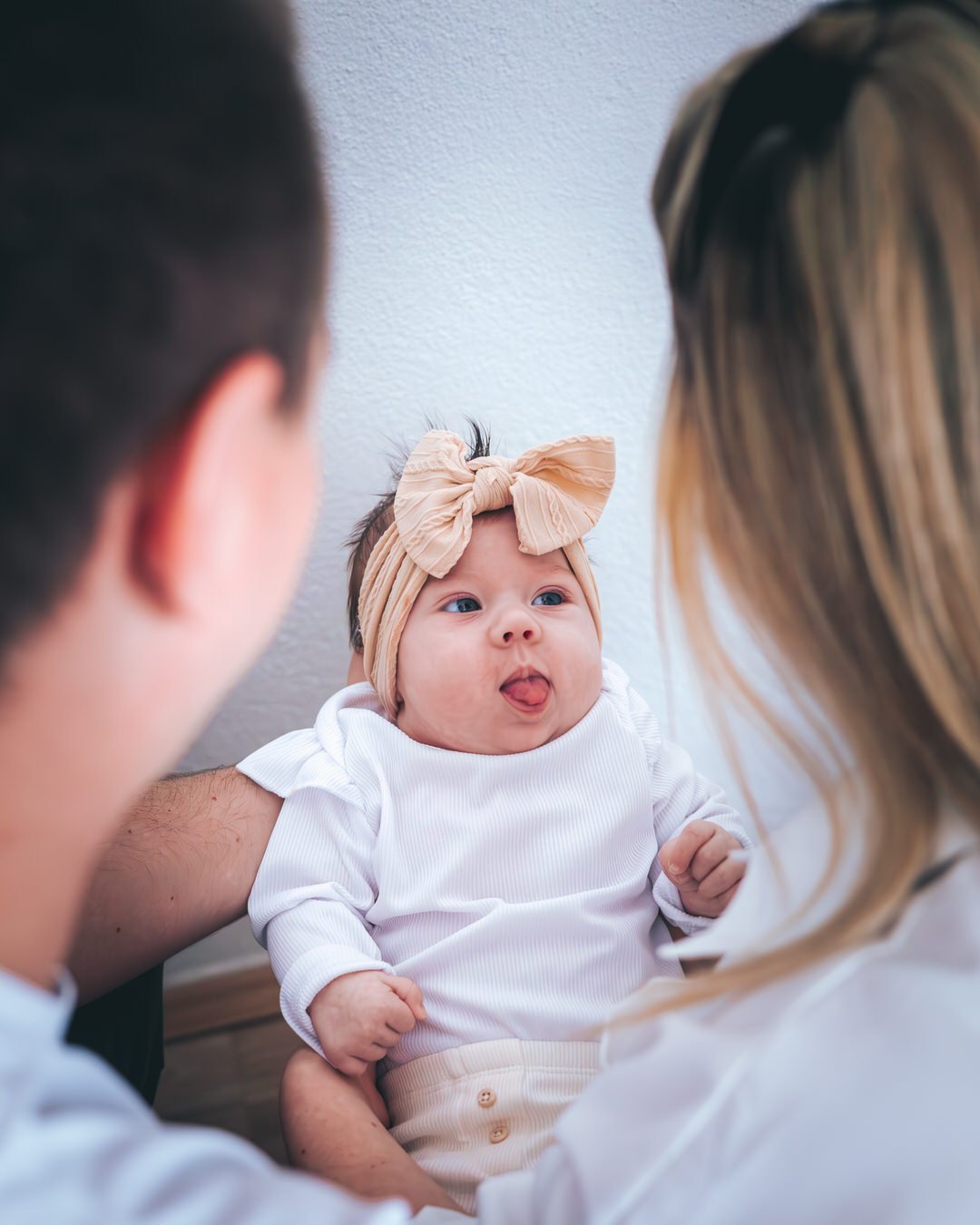A baby girl with a beige bow headband looking at her parents sticking out her tongue.