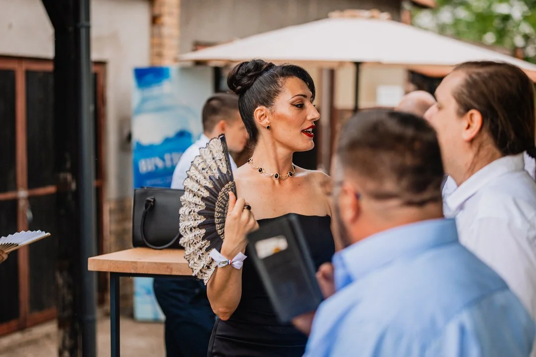 A woman wearing a black off-shoulder dress and jewelry, holding a decorative fan, engaging in conversation with men at a social gathering outdoors.