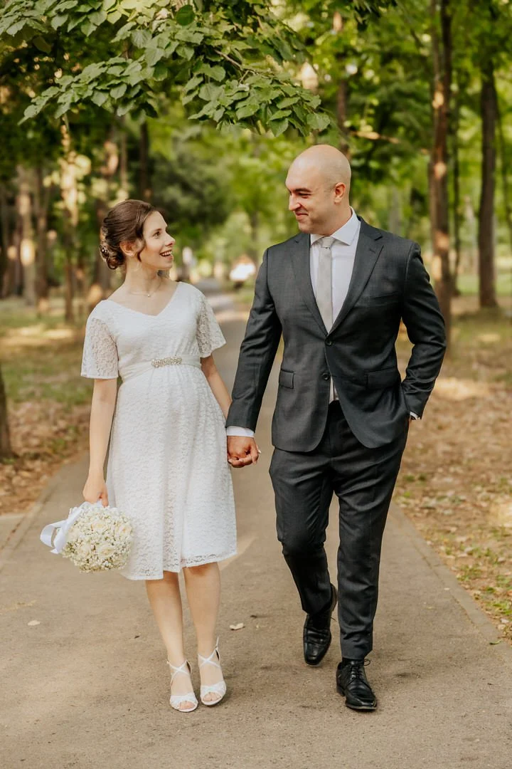 Bride and groom walking hand in hand along a pathway in Park Forest Adica, Vukovar, surrounded by lush green trees, with the bride holding a bouquet of white flowers.