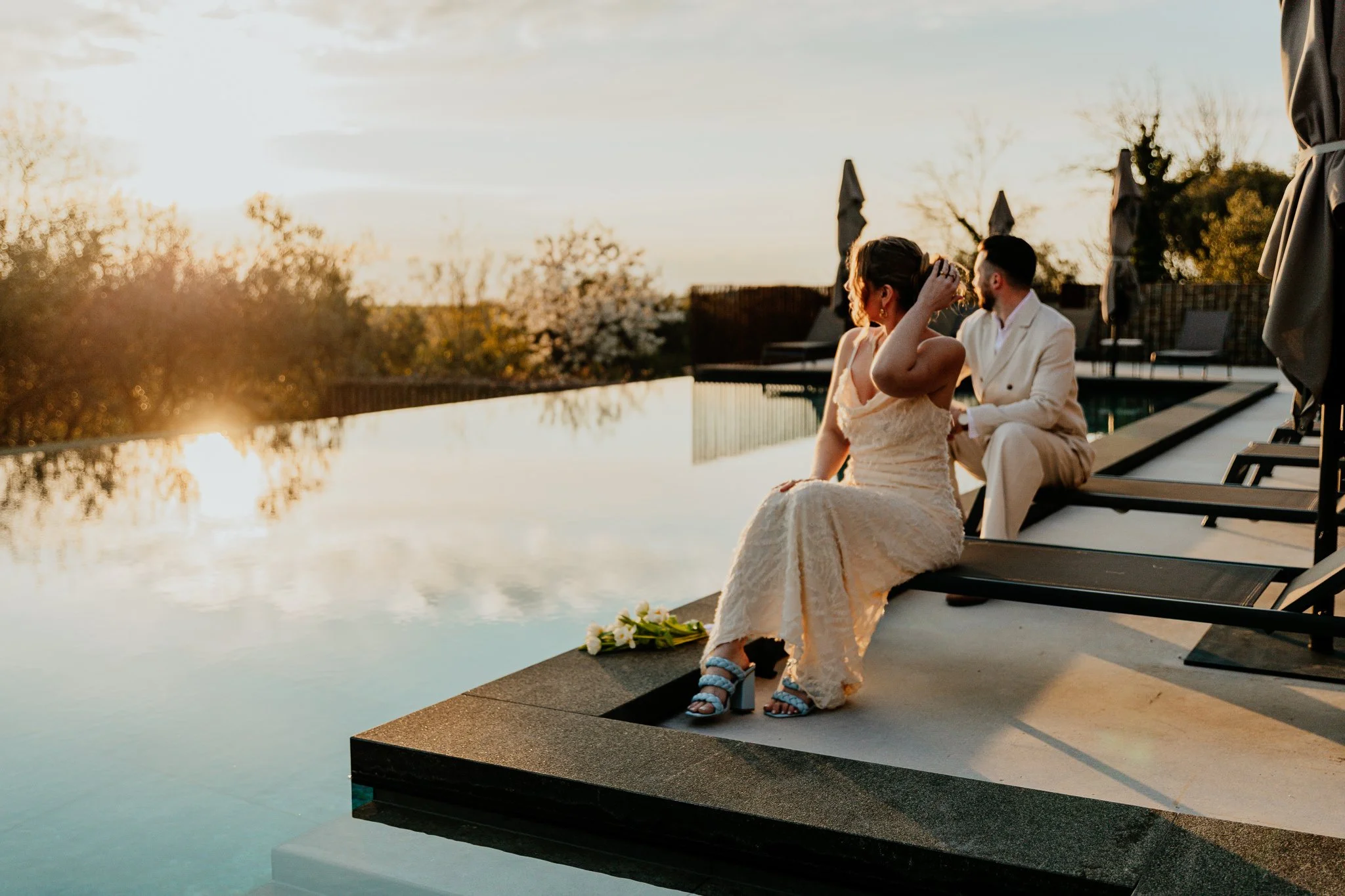 A woman in a beige dress and a man in a white suit sitting by a pool at sunset. The woman adjusts her hair, and the man looks towards her. Flowers are placed on the poolside.