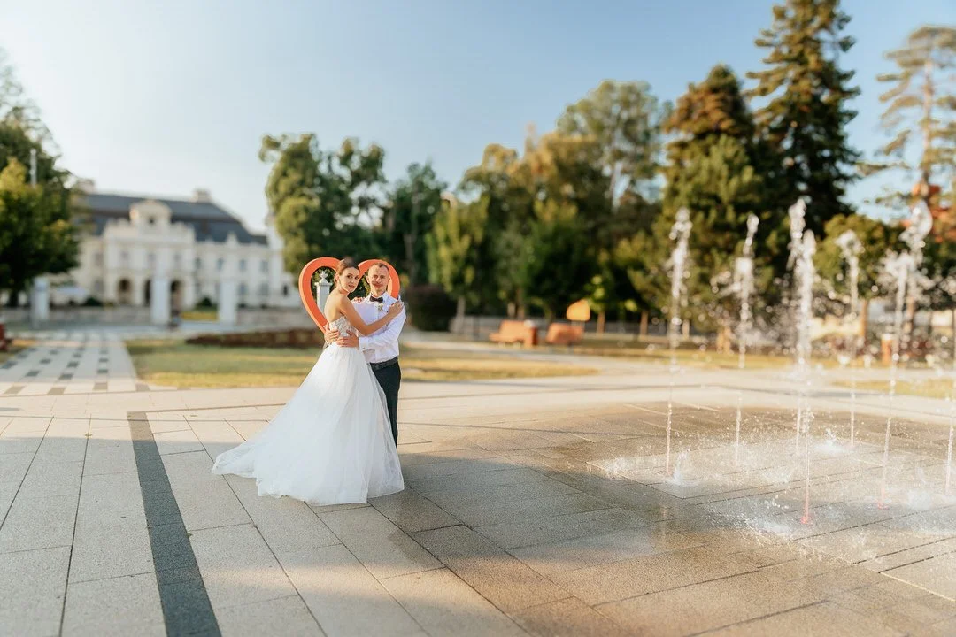 A bride and groom in wedding attire posing near a fountain in a park.