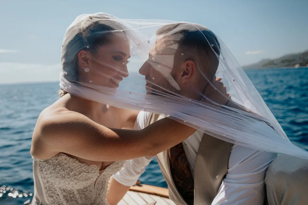 A bride and groom on a boat, with the bride's veil covering both of their faces as they smile at each other, across the ocean with a distant land visible.
