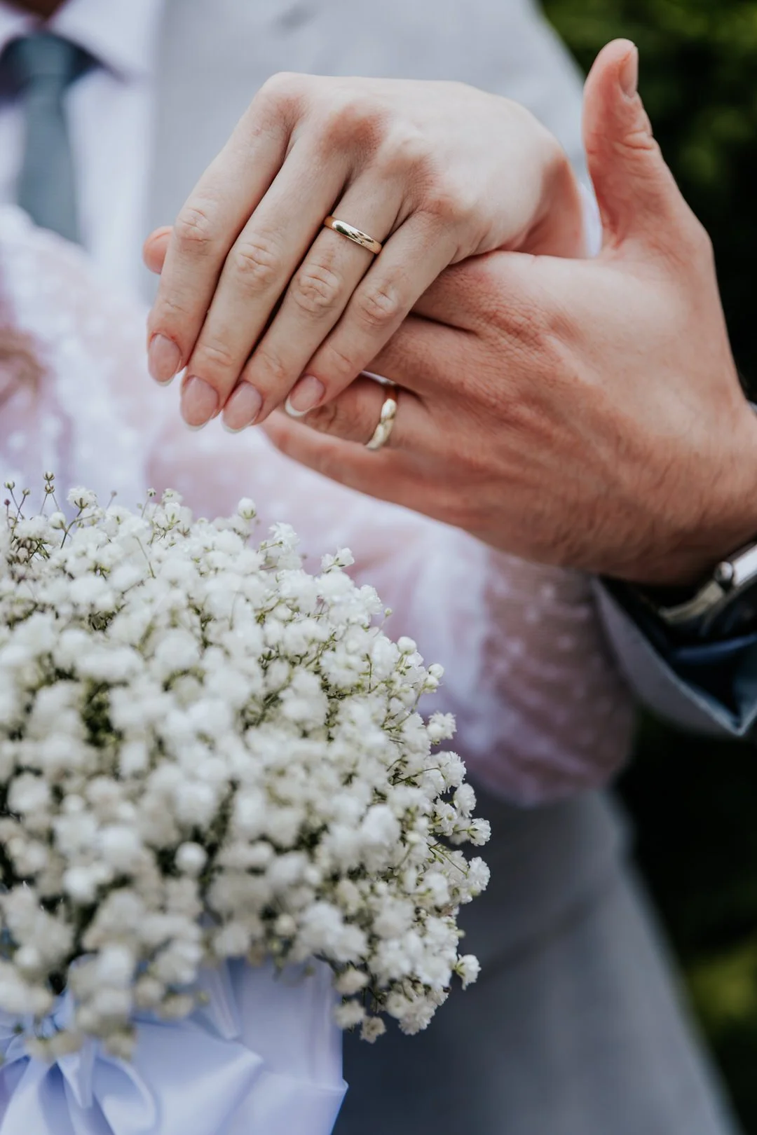 Close-up of a bride and groom's hands showing wedding rings, with a bouquet of white flowers in the foreground.