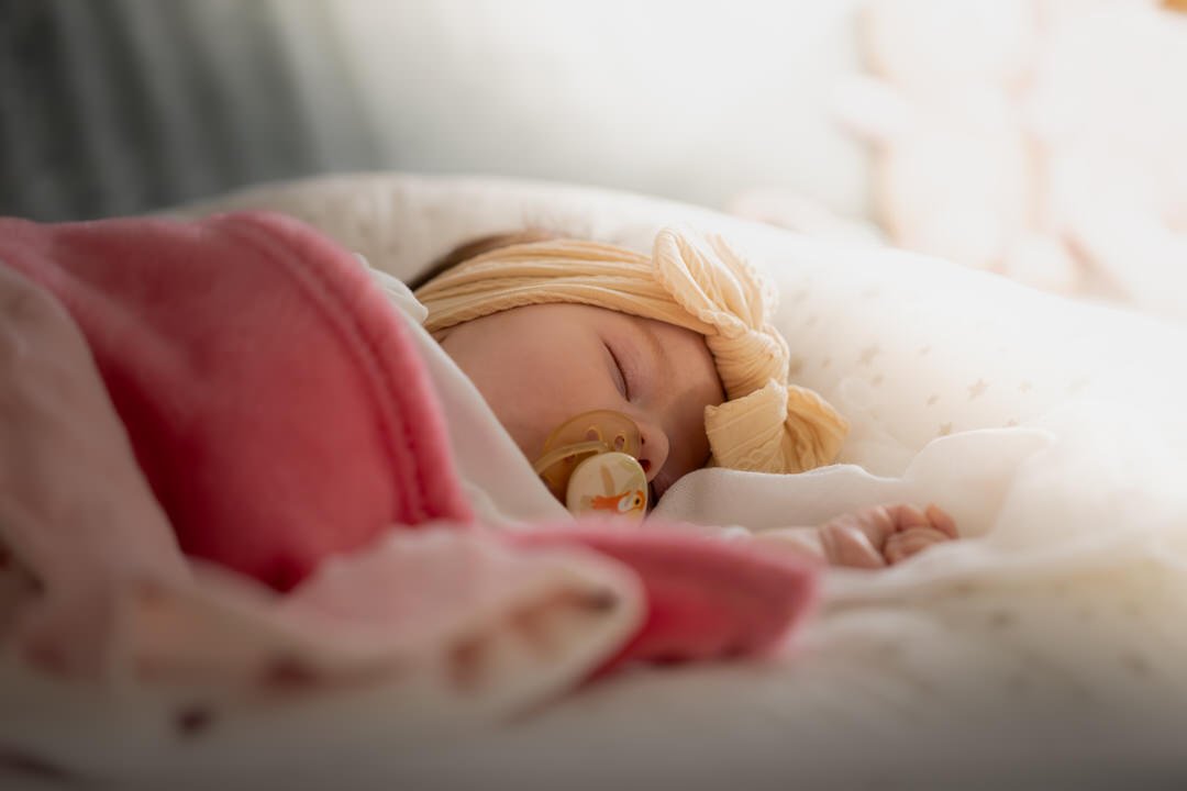 A baby with a pacifier sleeping on a bed, wearing a beige headband with a bow.