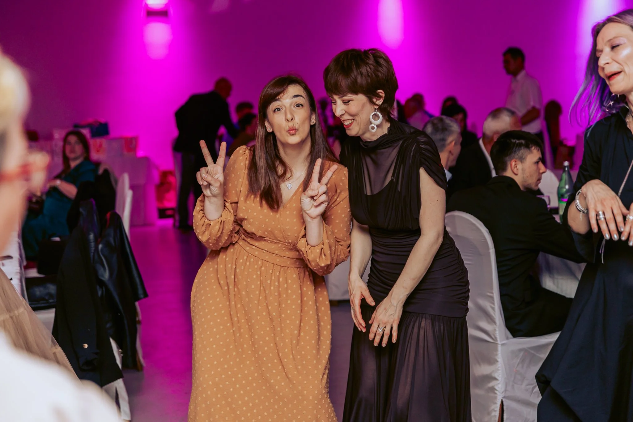 Two women at a party, one making a peace sign and the other smiling, with a pink-lit background and other guests seated and standing.